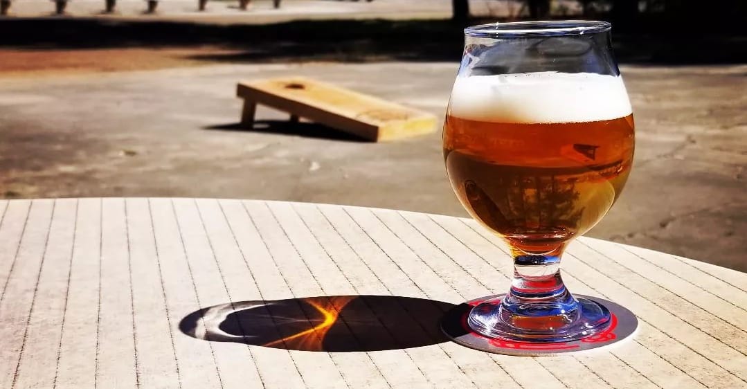 A glass of amber beer on a sunny patio table with a cornhole board in the background